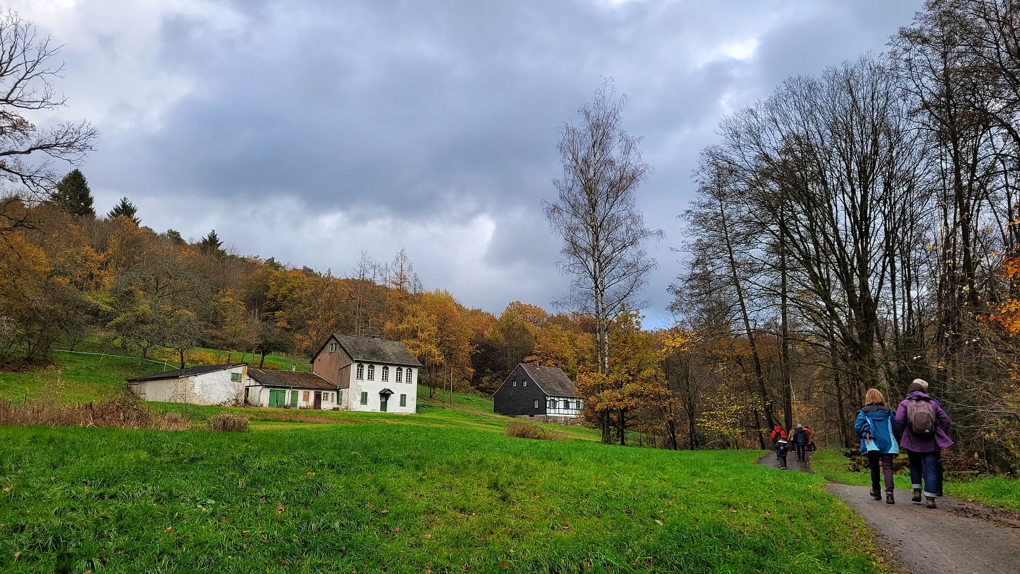 idyllische Landschaft mit einem Gehöft und einem Fachwerk-Schiefer-Haus; rechts vorne einige Wanderer