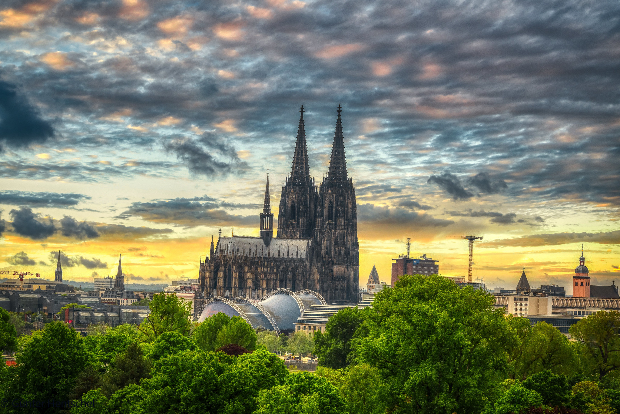 Kölner Dom vor dramatisch gelb-blau-grauem Himmel; vorne grüne Bäume